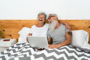 Happy senior couple having fun watching media on computer inside bed - Elderly people love and relationship concept photo