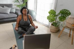 joven africano curvilíneo mujer haciendo yoga virtual aptitud clase con ordenador portátil a hogar - deporte bienestar personas estilo de vida concepto foto