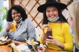 Multiracial friends having healthy lunch drinking fruits fresh smoothies in coffee brunch bar during corona virus outbreak photo