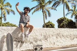 Young Latin man having fun taking selfie with mobile smartphone while listening music with headphones and boombox during summer vacations photo