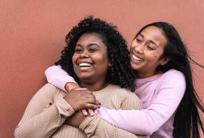 Happy Latin girls having fun embracing while standing against red wall - Young people lifestyle and friendship concept photo