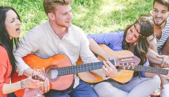 Group of friends playing guitars and singing while drinking red wine sitting on grass in a park outdoor - Happy people having fun together making a picnic in the garden - Friendship, lifestyle concept photo