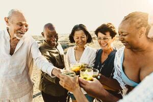 Happy multiracial senior friends having fun drinking and toasting mojitos on the beach during sunset time - Elderly people enjoying summer holidays photo