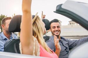 Happy friends having fun in convertible car at sunset in vacation - Young rich people making party and dancing in auto cabriolet during their road trip - Friendship, travel, youth lifestyle concept photo