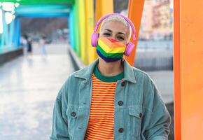 Young woman wearing gay pride mask while listening to music with headphones outdoor - Gender equality and technology concept photo