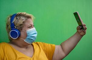 Plus size woman taking selfie with mobile smartphone and listening to music with wireless headphones while wearing face protective mask to avoid corona virus infection - Health and technology concept photo