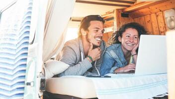 Happy couple watching videos on their computer inside minivan at sunset - Travel couple using laptop during their journey on a vintage mini van with wood interior - Vacation, love, technology concepts photo