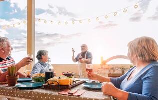 Happy senior friends having fun dining  at barbecue in terrace outdoor - Older people enjoying together drinking red wine and grill meat - Friendship and elderly lifestyle concept photo