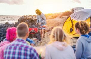Group of happy friends camping with tent on the beach and preparing a barbecue dinner - Young people having fun making bbq and drinking beer next to ocean - Vacation, youth, travel lifestyle concept photo