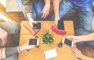 Top view of a group of hipster friends sitting in a bar cafe using mobile smart phone - New young generation trend - concept of people merged with phone technology - Focus on male hand on left photo