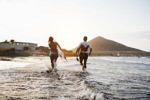 Happy surfers running in the water at sunset time - Young couple having fun surfing in ocean - Extreme sport and youth culture lifestyle concept photo