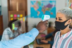 Young people in co-working creative space checkup temperature with digital thermometer while wearing face mask and keeping social distance to avoid corona virus spread photo