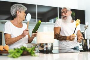 Happy senior couple having fun cooking together at home - Elderly people preparing lunch in modern kitchen - Retired lifestyle family time and food nutrition concept photo