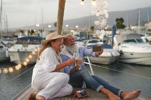 Senior couple toasting champagne while taking selfie on sailboat vacation - Happy mature people having fun celebrating wedding anniversary on boat trip - Love relationship and travel lifestyle concept photo