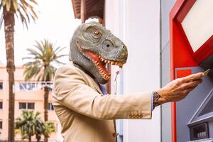 Senior man wearing t-rex dinosaur mask withdraw money from bank cash machine with debit card - Surreal image of half human and animal - Absurd and crazy concept of ATM advertise photo