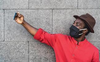 African fashion man taking selfie with smartphone and wearing face black mask to avoid corona virus spread - People health care and technology concept photo