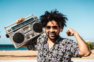 contento latín hombre teniendo divertido escuchando a música con inalámbrico auriculares y Clásico boombox en el playa durante verano hora foto