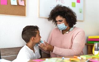 Female teacher giving face protective mask to student in preschool classroom during corona virus pandemic - Healthcare and education concept photo