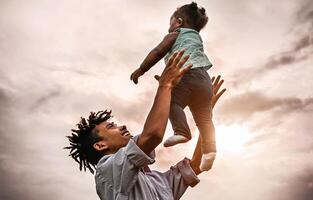 Happy father playing with his baby daughter during sunset time - Afro family having fun outdoor - Concept of child, happiness and parenthood photo