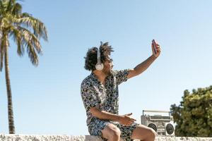 Young man having fun taking selfie with mobile smartphone while listening music with headphones and vintage boombox during summer vacations photo