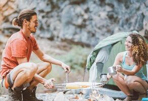 excursionista Pareja cámping en rock montañas con un tienda - trepador personas Cocinando y Bebiendo caliente té siguiente a hoguera - viajar, extremo deporte, salvaje vida concepto foto