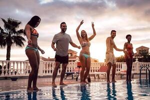 Group of happy friends dancing at pool party at sunset - Young millennial people having fun in a tropical luxury resort - Vacation, summer holidays and youth lifestyle concept photo
