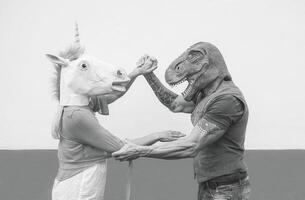 Crazy couple dancing and wearing dinosaur and unicorn mask - Senior trendy people having fun masked at carnival parade - Absurd, eccentric, surreal, fest and funny masquerade concept photo