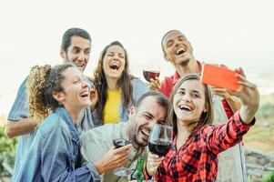 grupo de contento amigos tomando selfie utilizando móvil inteligente teléfono cámara - joven personas teniendo divertido un Bebiendo rojo vino al aire libre - amistad, tecnología, juventud estilo de vida concepto foto