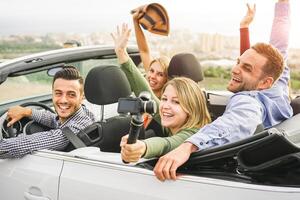 Happy friends taking photos with selfie stick camera in convertible car in vacation - Young people having fun in cabriolet auto during their road trip - Friendship, travel, youth lifestyle concept