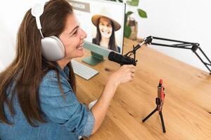 Mature woman recording a podcast using microphone and laptop while streaming on social network with mobile smartphone from her home studio photo