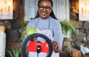 African senior woman preparing food recipe while streaming online with mobile smartphone cam for web kitchen masterclass channel photo