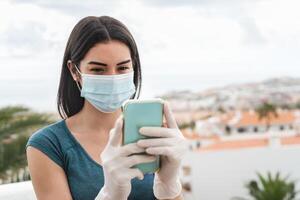 Young woman wearing surgical face mask using mobile smartphone o home terrace - Girl in quarantine Having fun with new technology social media apps - Corona virus confinement and Youth people concept photo
