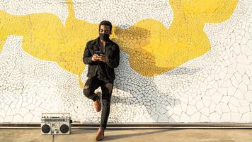 Young Afro man using mobile smartphone while standing against urban wall and listening music with headphones and vintage boombox photo