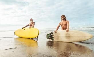 Happy fit friends having fun surfing on tropical ocean - Surfers father and son doing stretching surf exercises - Sporty people lifestyle and extreme sport concept photo