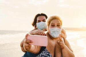 Happy plus size women taking selfie with mobile smartphone on the beach - Overweight friends having fun on vacation during corona virus pandemic - Healthcare and technology concept photo