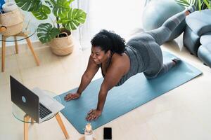 joven africano curvilíneo mujer haciendo pilates virtual aptitud clase con ordenador portátil a hogar - deporte bienestar personas estilo de vida concepto foto