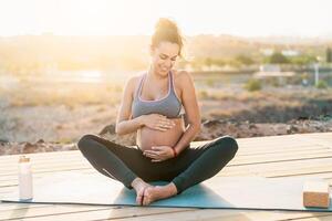 Young pregnant woman massaging the belly while doing yoga meditation outdoor - Health lifestyle and maternity concept photo