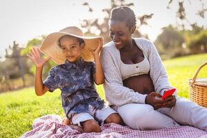 Happy African pregnant mother spending time with his son doing a picnic during weekend in public park - Maternity and parents lifestyle concept photo