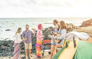 Group of happy friends celebrating drinking beer and playing guitar while camping with tent - Surfers people having fun listening music at barbecue picnic outdoor - Vacation, travel lifestyle concept photo