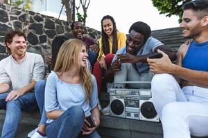 Young multiracial friends having fun listening music with vintage boombox stereo while sitting on city urban stairs - Youth millennial lifestyle concept photo