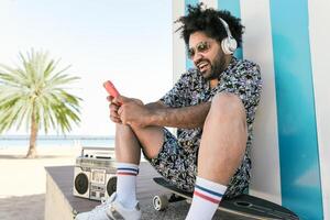 afro latín hombre teniendo divertido con móvil teléfono inteligente y escuchando música con auriculares y Clásico boombox estéreo en tropical playa durante vacaciones hora foto