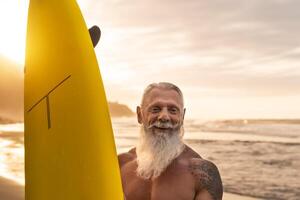 Happy fit senior having fun surfing at sunset time - Sporty bearded man training with surfboard on the beach - Elderly healthy people lifestyle and extreme sport concept photo