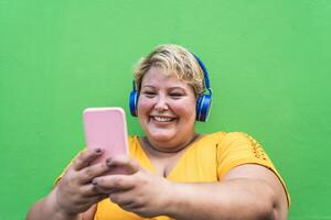 Happy plus size woman taking selfie with mobile smartphone and listening music with wireless headphones - Confident overweight people and technology concept photo