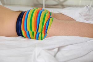 Close-up young adult gay man, non binary person, sleeping on top of white bed sheets in rainbow LGBT pants photo