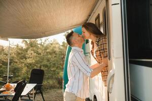 un casado Pareja soportes siguiente a su casa rodante a puesta de sol en el bosque y Beso foto