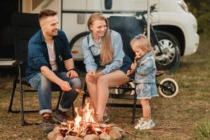 un familia cocineros salchichas en un hoguera cerca su casa rodante en el bosque foto