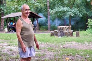 sonriente mayor hombre pensionista en un camiseta y pantalones cortos en un bosque en un picnic foto