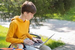 Thoughtful teenager boy working on laptop. Holding and using a laptop for networking on a sunny spring day, outdoors. photo