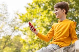 un joven chico es teniendo un vídeo llamada al aire libre, participación un teléfono inteligente en su manos. foto