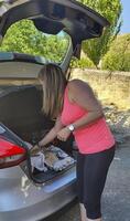 joven rubia mujer, en Deportes ropa, preparando almuerzo en el maletero de un coche antes de el caminar. foto